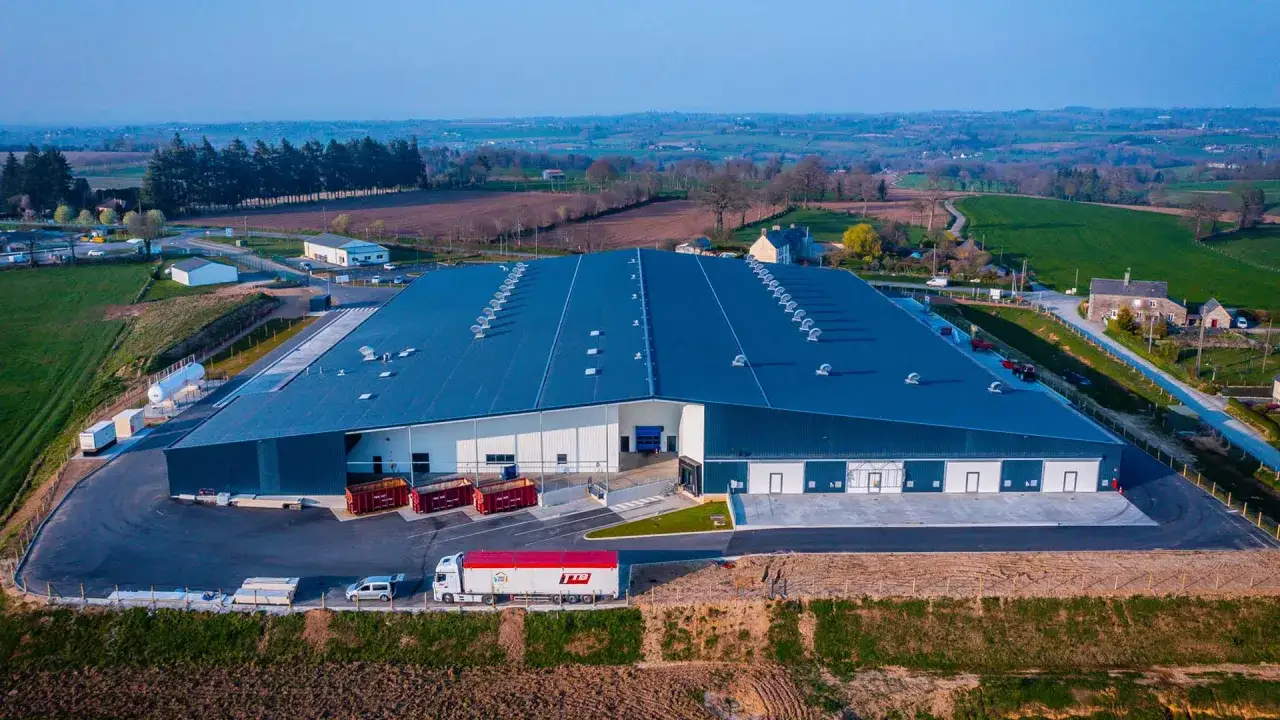 Duży, nowoczesny magazyn z niebieskim dachem i otoczeniem pól i drzew.