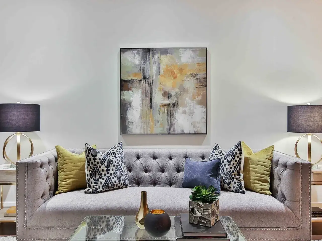 Id&eacute;e pour comment d&eacute;corer un grand mur de salon : un tableau abstrait moderne au-dessus d'un canap&eacute; gris capitonn&eacute;, flanqu&eacute; de lampes et coussins color&eacute;s.