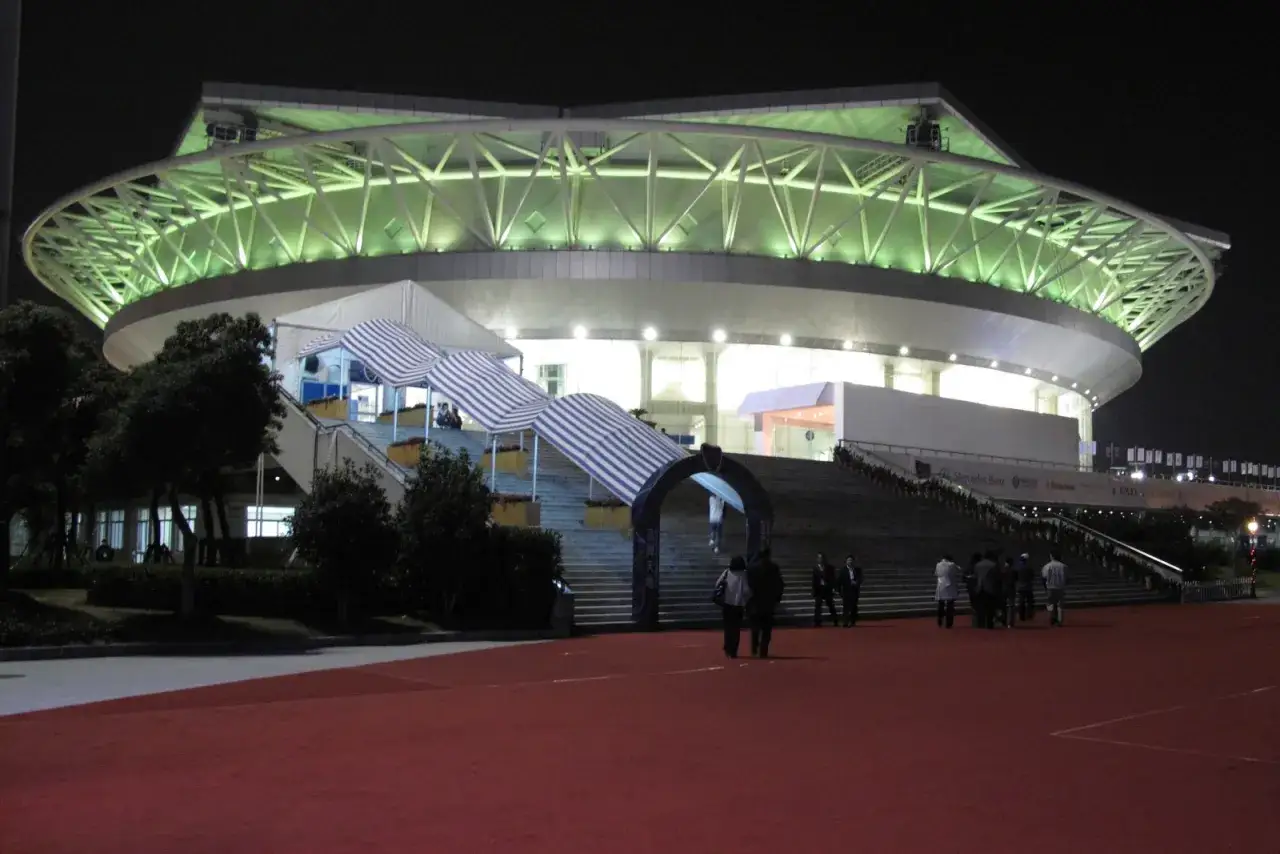 El estadio Master Mil Shanghai, iluminado de noche, con una estructura de techo verde y escaleras de acceso cubiertas.
