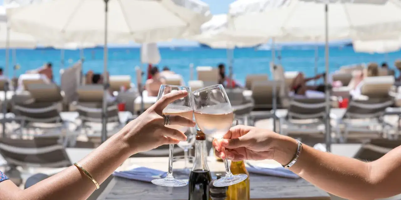 Deux mains portent des verres de ros&eacute; pour un toast, avec la mer bleue et des parasols en arri&egrave;re-plan. Ambiance estivale dans un bar de plage &agrave; Cannes.
