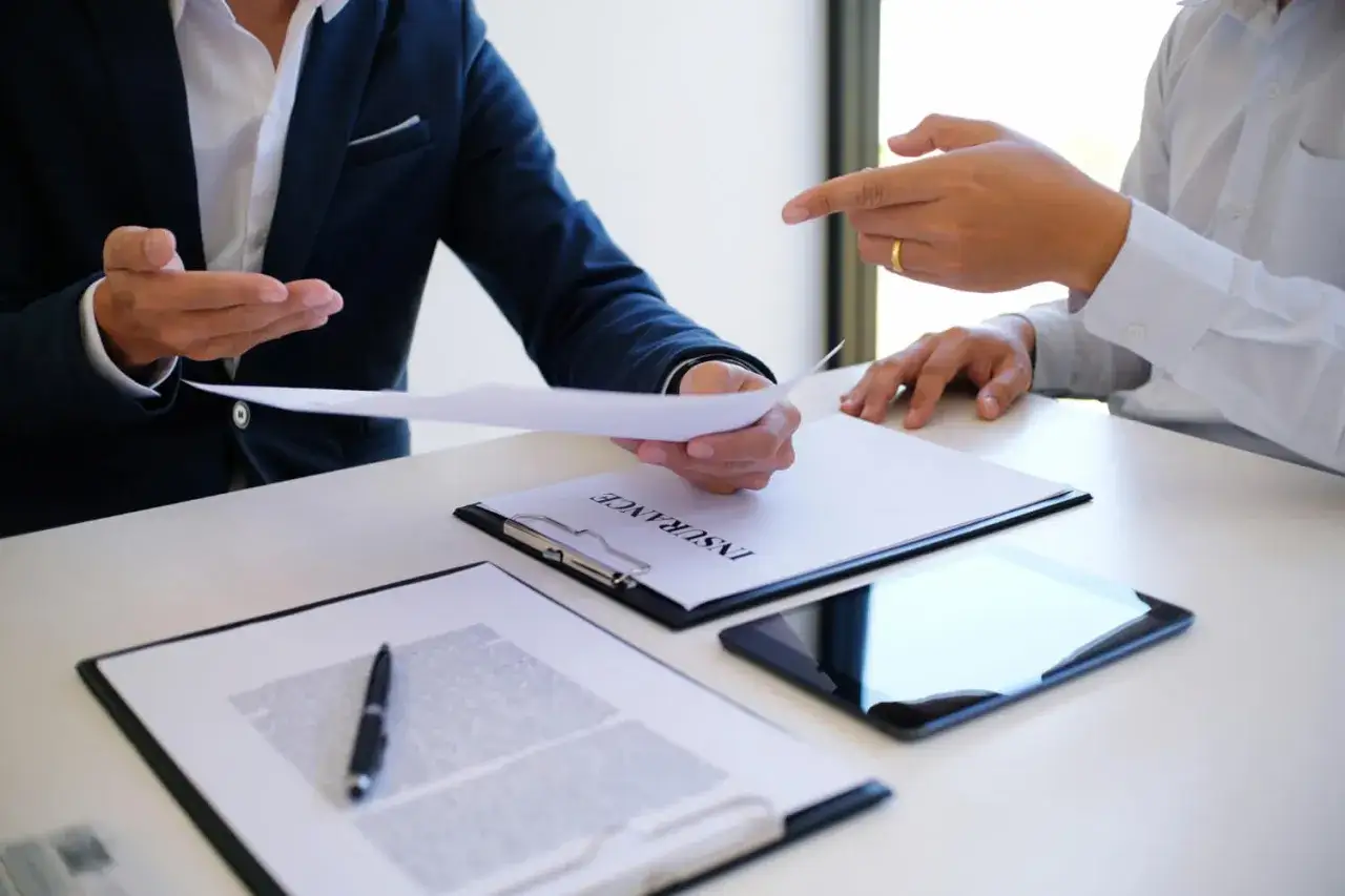 Dos hombres discuten documentos en una mesa. Uno entrega un papel al otro, quien se&ntilde;ala. Posiblemente brokers hipotecarios revisando un contrato.