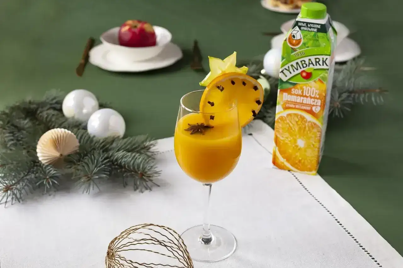 Pomarańczowy drink z gwiazdką anyżu i plasterkiem pomarańczy, jakby zrobiony z soku Tymbark. Obok kartonik soku, gałązki choinki i ozdoby.