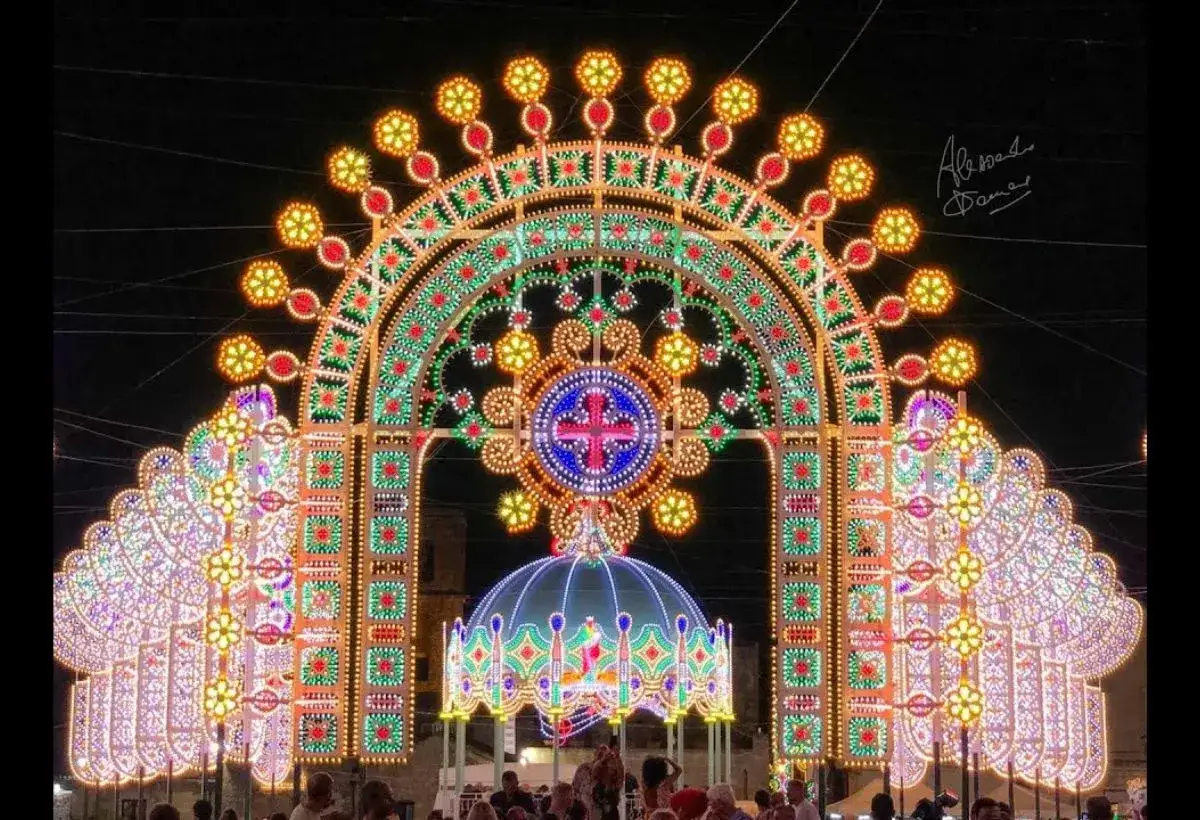 Un imponente arco di luci illumina la notte, un vero paese delle luminarie puglia. Dettagli floreali e un rosone centrale con croce blu e rossa dominano la scena.