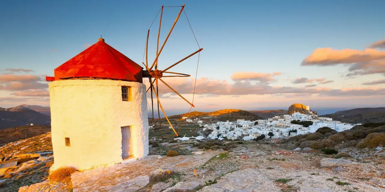 Väderkvarn med rött tak på Amorgos, Grekland, med en vit by i bakgrunden vid solnedgången.