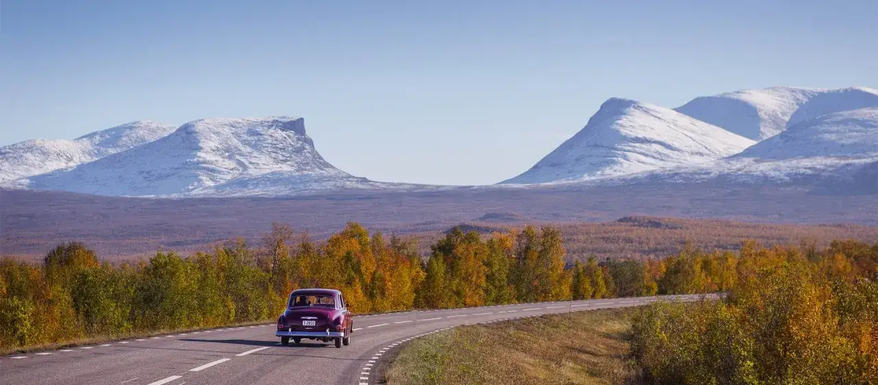 En lila veteranbil kör på en väg genom höstlandskap med snöklädda berg i bakgrunden. En påminnelse om att vissa saker, som mest pålitliga bilar, håller för evigt.