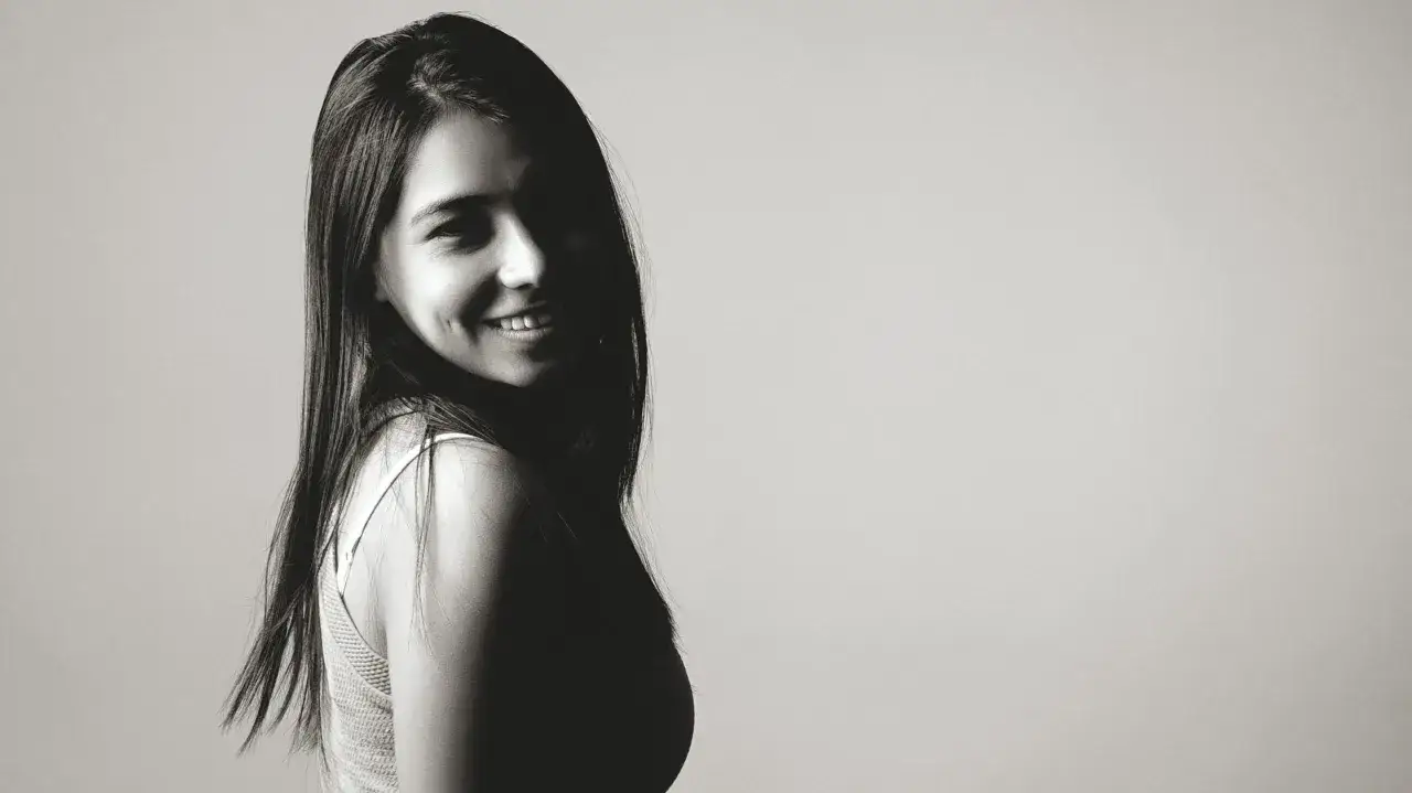 Foto ritratto in bianco e nero di una giovane donna con lunghi capelli scuri che sorride timidamente, illuminata da un lato.