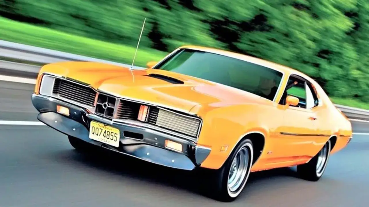 Pomarańczowy Mercury Cougar z lat 70. pędzi po drodze, otoczony rozmytą zielenią.