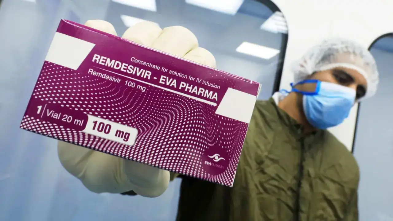 Ręka w rękawiczce trzyma pudełko leku Remdesivir 100 mg. W tle osoba w masce i czepku.