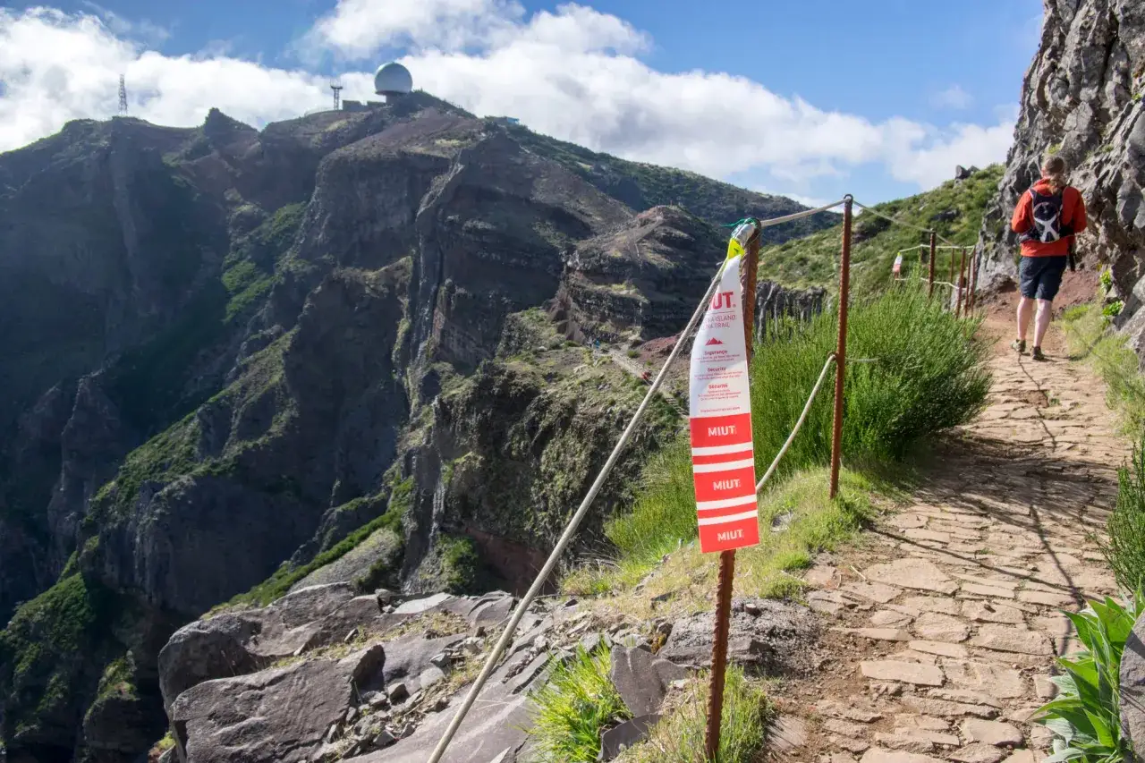 Wanderer auf einem steinigen Pfad auf Madeira, vorbei an einem roten MIUT-Schild. Im Hintergrund dramatische Berge und eine Kuppel.
