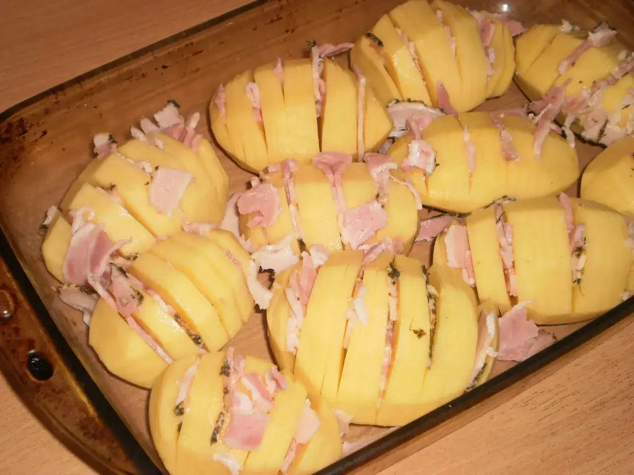 Ziemniaki hasselback z boczkiem i serem, gotowe do pieczenia w szklanym naczyniu.
