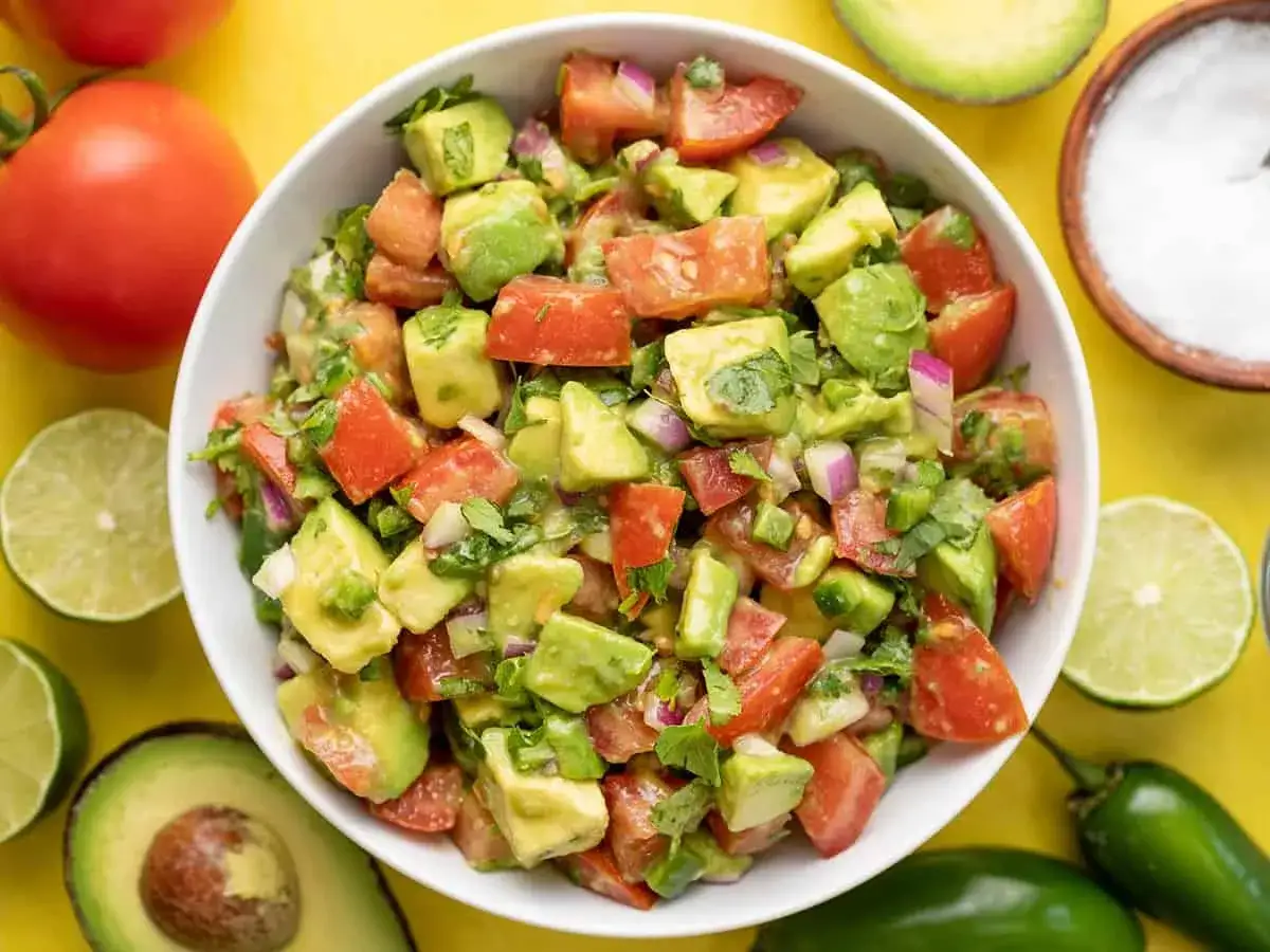 Frischer Avocado Tomaten Salat, garniert mit Koriander und Zwiebeln, umgeben von Limetten, Tomaten und Jalapeños.