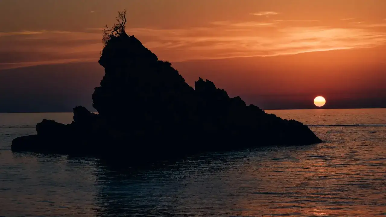 Tramonto infuocato sulle isole calabresi. Una roccia scura si staglia contro il cielo arancione, con un albero solitario in cima.