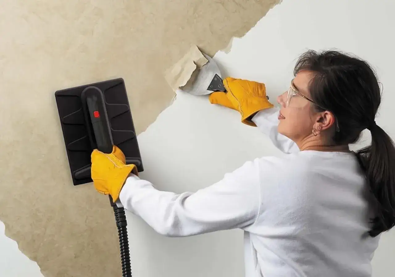 Une femme, portant des gants jaunes, utilise un appareil pour d&eacute;coller du papier peint ancien d'un mur.