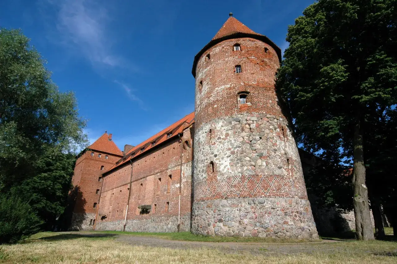 Zamek w Bytowie, ceglana twierdza z kamienną podstawą, góruje nad zielenią.