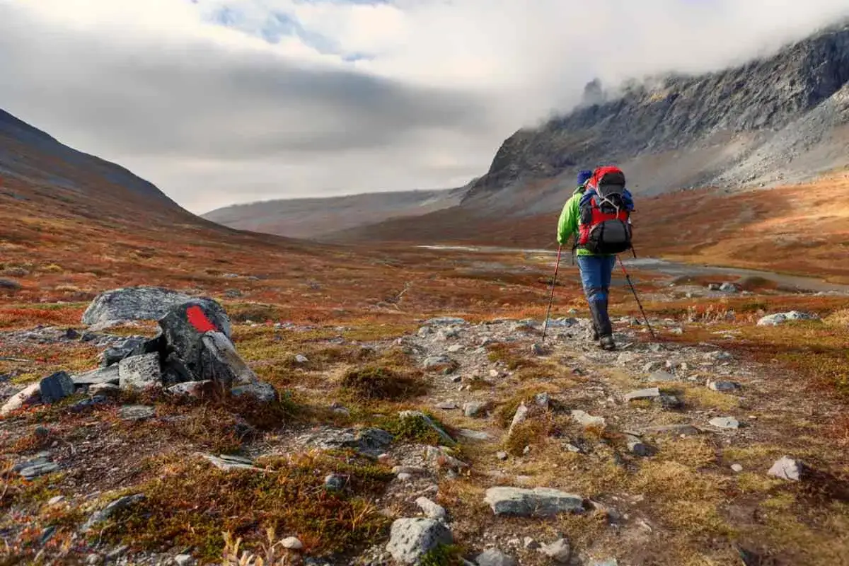 Vandrare med ryggs&auml;ck och stavar p&aring; en stig i fj&auml;llen, med en r&ouml;d markering p&aring; en sten. En vy som p&aring;minner om vandringen mellan Kvikkjokk och Saltoluokta.