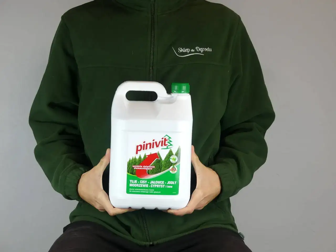 Osoba w zielonej bluzie trzyma białą bańkę z preparatem **pinivit** do nawożenia iglaków.