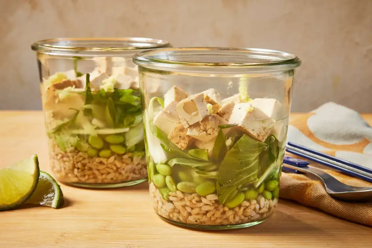 Deux bocaux de soupe japonaise avec riz, edamame, tofu et légumes verts. Des quartiers de citron vert sont à côté.