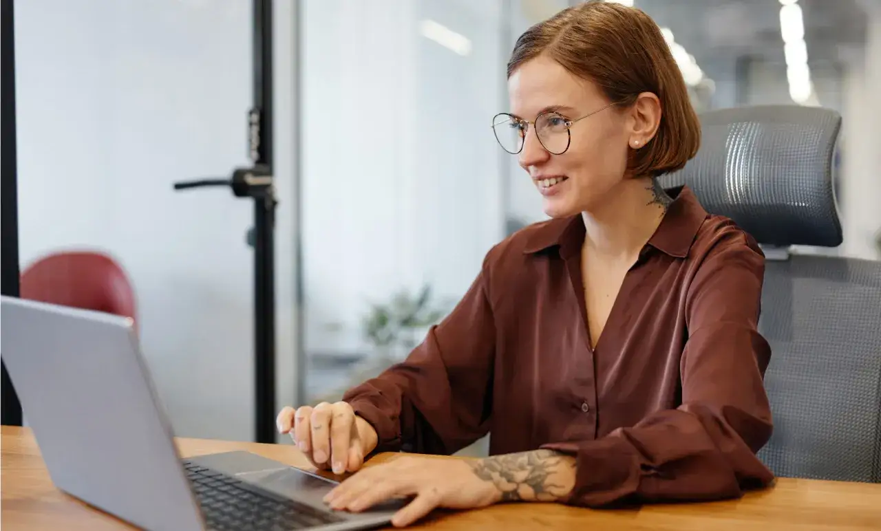 Kvinna med glas&ouml;gon och tatueringar ler vid sin laptop. Hon kanske k&auml;nner sig d&aring;lig p&aring; sitt jobb, men ser &auml;nd&aring; positiv ut.