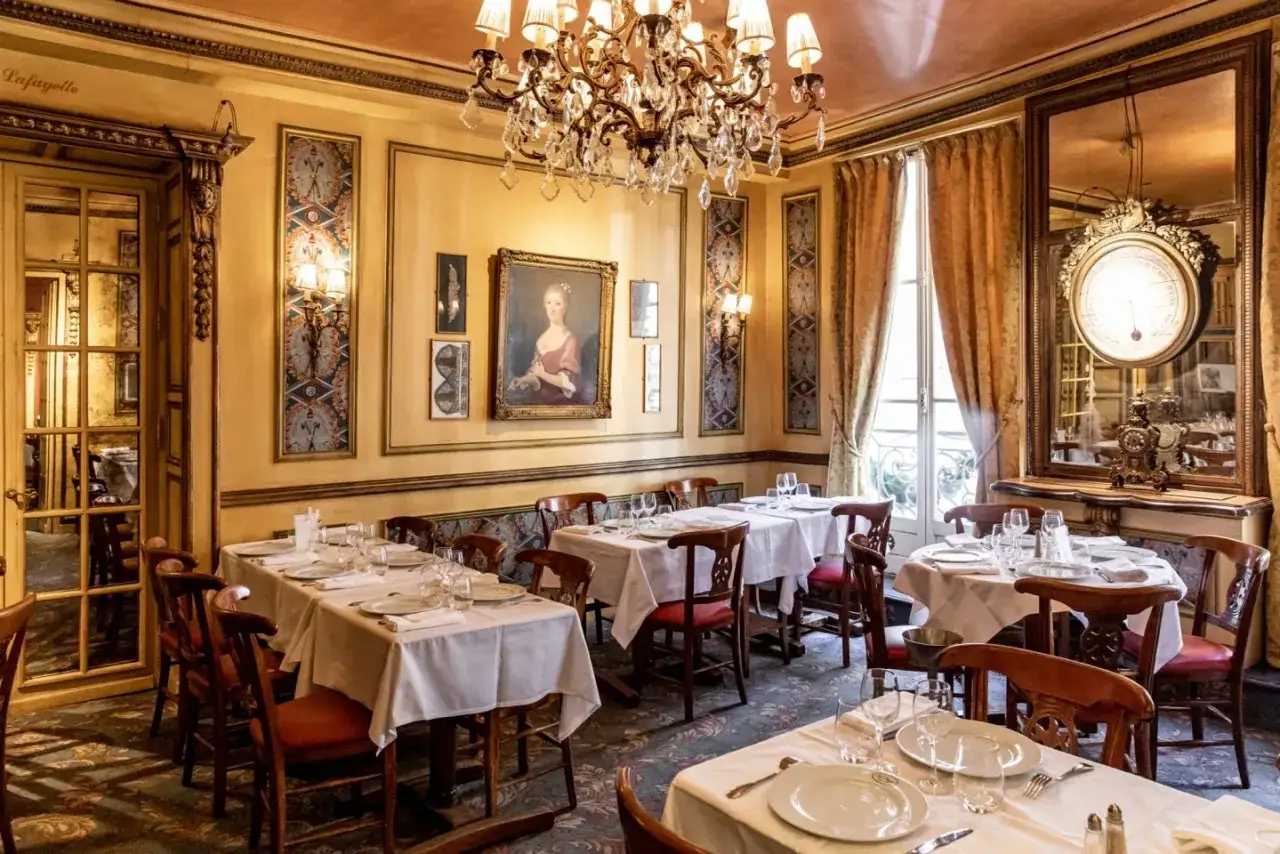 Interno elegante di uno dei **locali storici di Parigi**, con tavoli apparecchiati, un grande lampadario e un ritratto d'epoca.