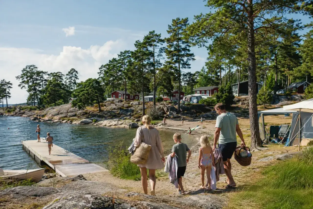Familj p&aring; v&auml;g mot vattnet f&ouml;r en h&auml;rlig sommarsemester med barn. Sol, bad och campingliv vid en svensk sj&ouml;.
