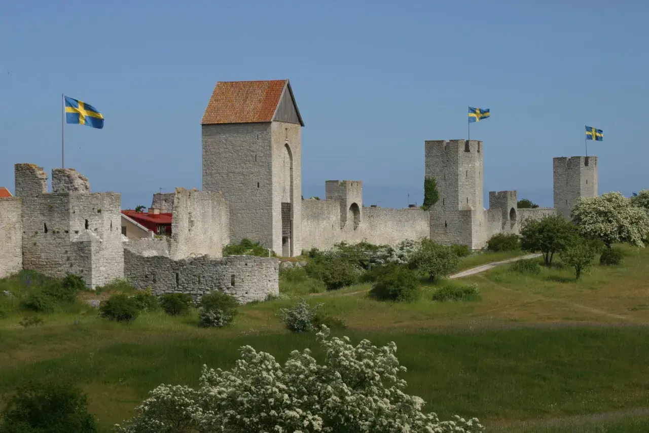 Visbys ringmur, en p&aring;minnelse om stadens rika visby historia, reser sig mot en klarbl&aring; himmel. Svenska flaggor vajar stolt fr&aring;n tornen.