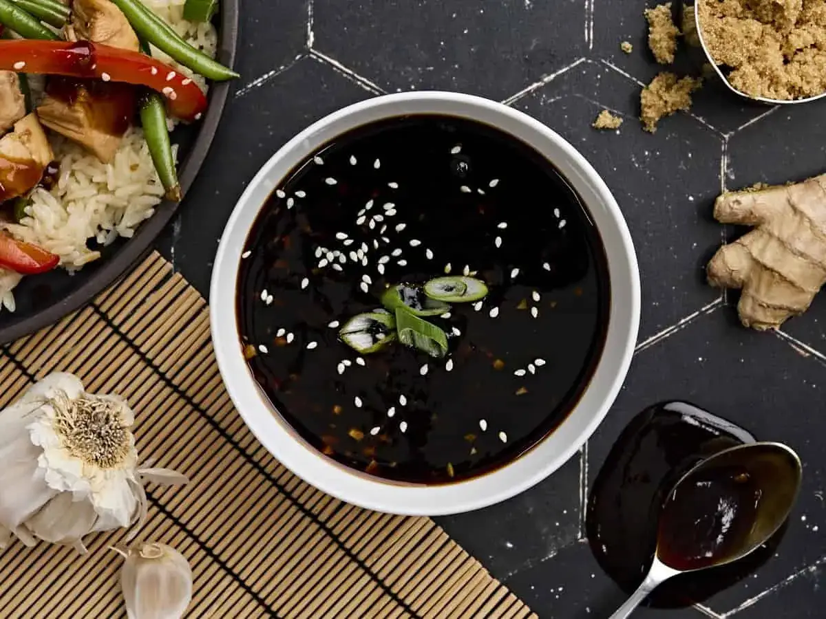 En skål med mörk teriyakisås, toppad med sesamfrön och salladslök. Ingredienser som ingefära och vitlök antyder ett utsökt teriyakisås recept.