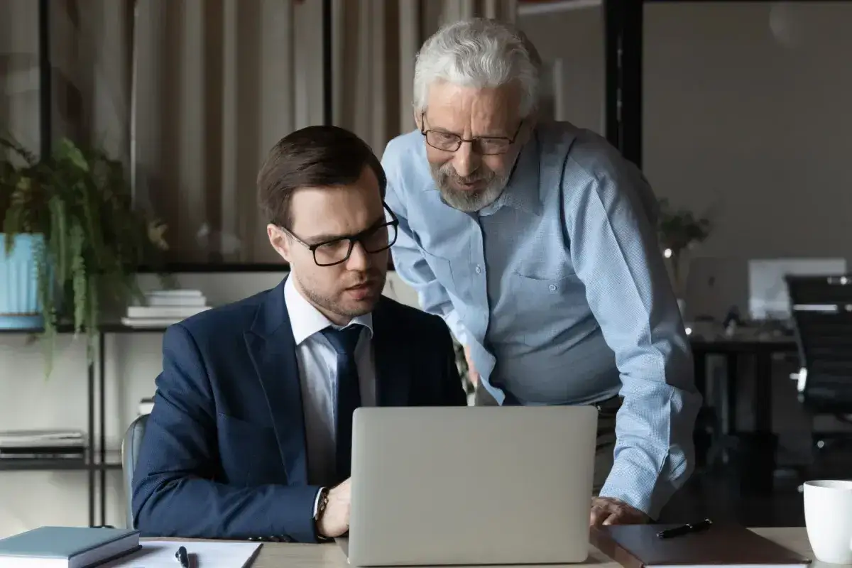 Dwóch mężczyzn przy laptopie, młodszy w garniturze, starszy pochyla się nad nim. Rozmawiają o czymś, co może być związane z ich pracą lub dodatkiem stażowym.