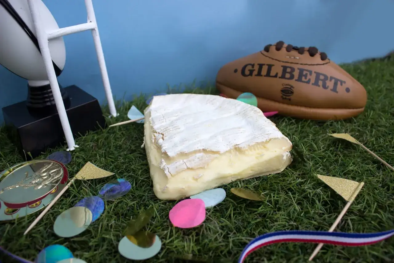 Un morceau de brie de Nangis, pr&ecirc;t &agrave; &ecirc;tre d&eacute;gust&eacute;, sur fond d'herbe avec un ballon de rugby et des d&eacute;corations festives.