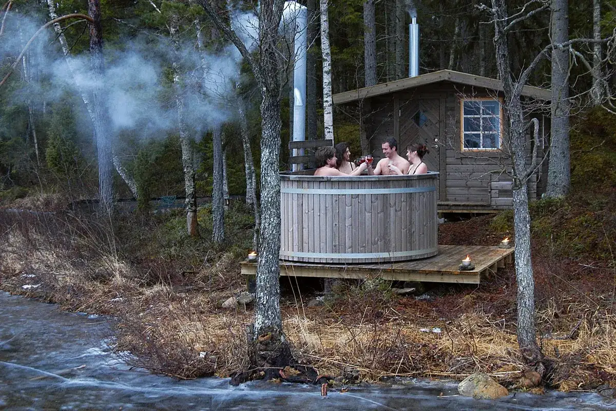 Fyra vänner njuter av en badtunna utomhus, med en rökig skorsten som vittnar om att de valt att bygga badtunna för avkoppling i skogen.