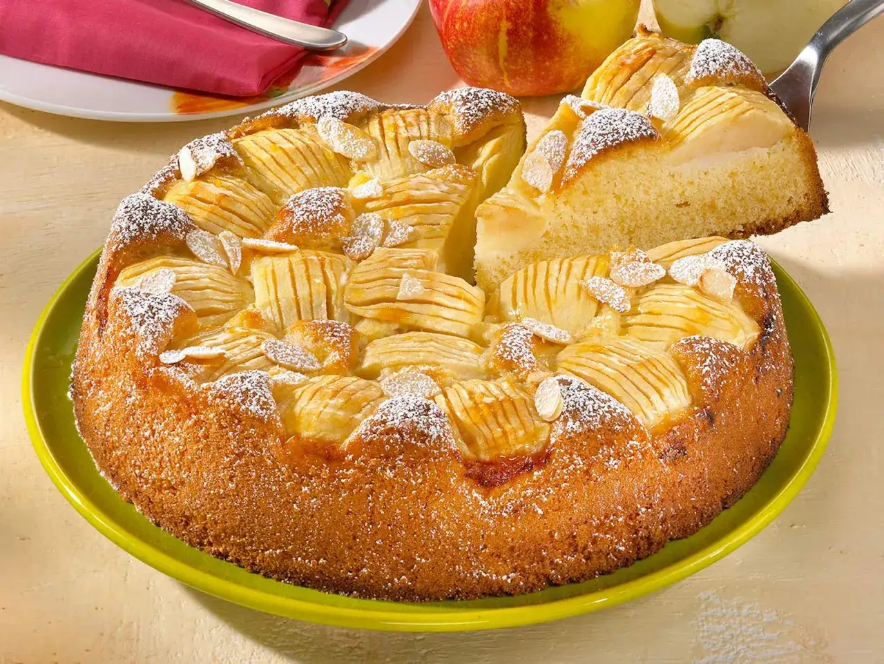 Ein saftiger, versunkener Apfelkuchen mit Mandelsplittern und Puderzucker bestreut, ein St&uuml;ck wird gerade herausgehoben.