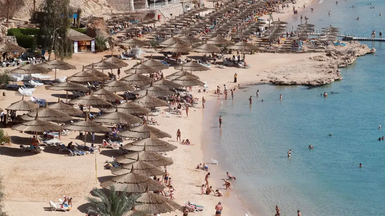 Tłum ludzi na plaży w Sharm el Sheikh, pod parasolami, w wodzie. Wiele osób wypoczywa, co sugeruje, że czy w Sharm el Sheikh jest bezpiecznie.