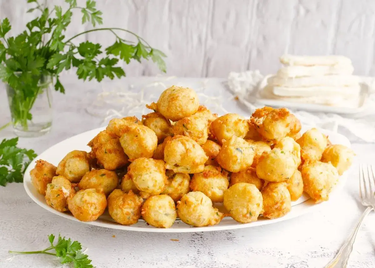 Montón de buñuelos de bacalao dorados y esponjosos en un plato blanco, decorados con perejil fresco.