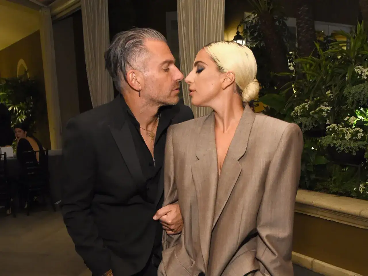 Lady Gaga i Christian Carino w intymnym uścisku, otoczeni zielenią.