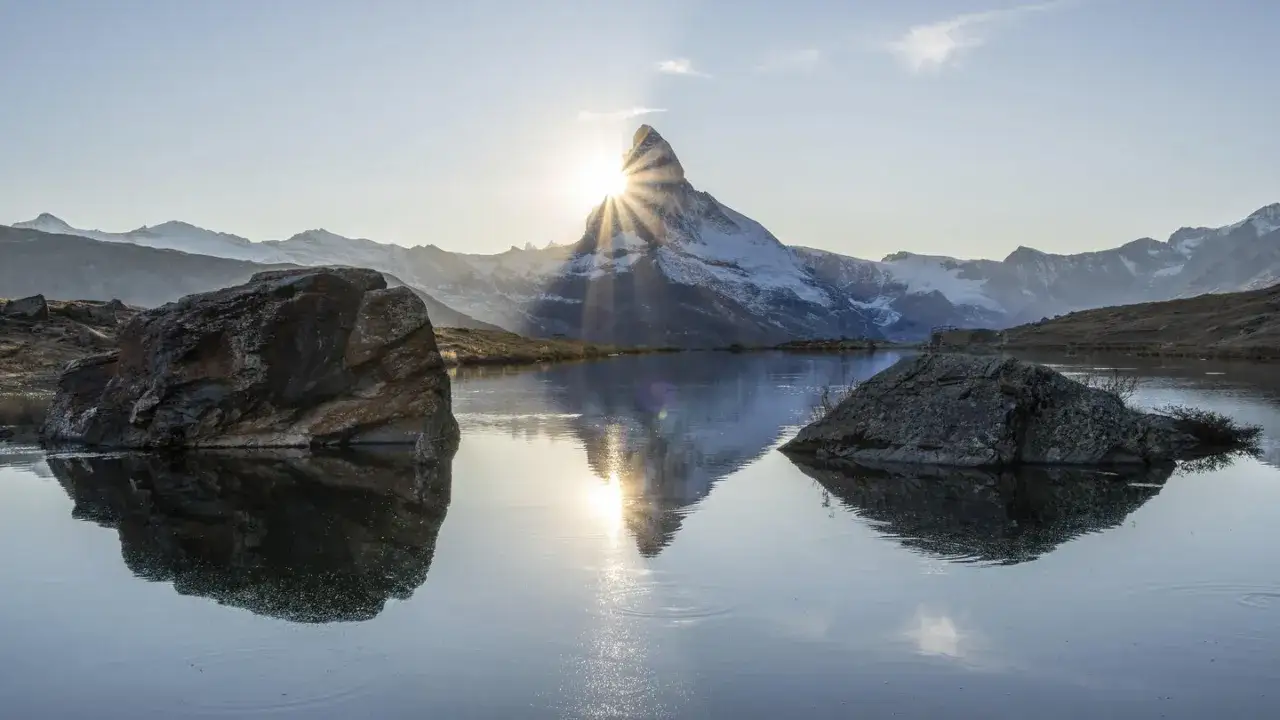 Sonnenstrahlen brechen durch das Matterhorn, wie in Psalm 102 beschrieben. Felsen spiegeln sich im ruhigen Bergsee.