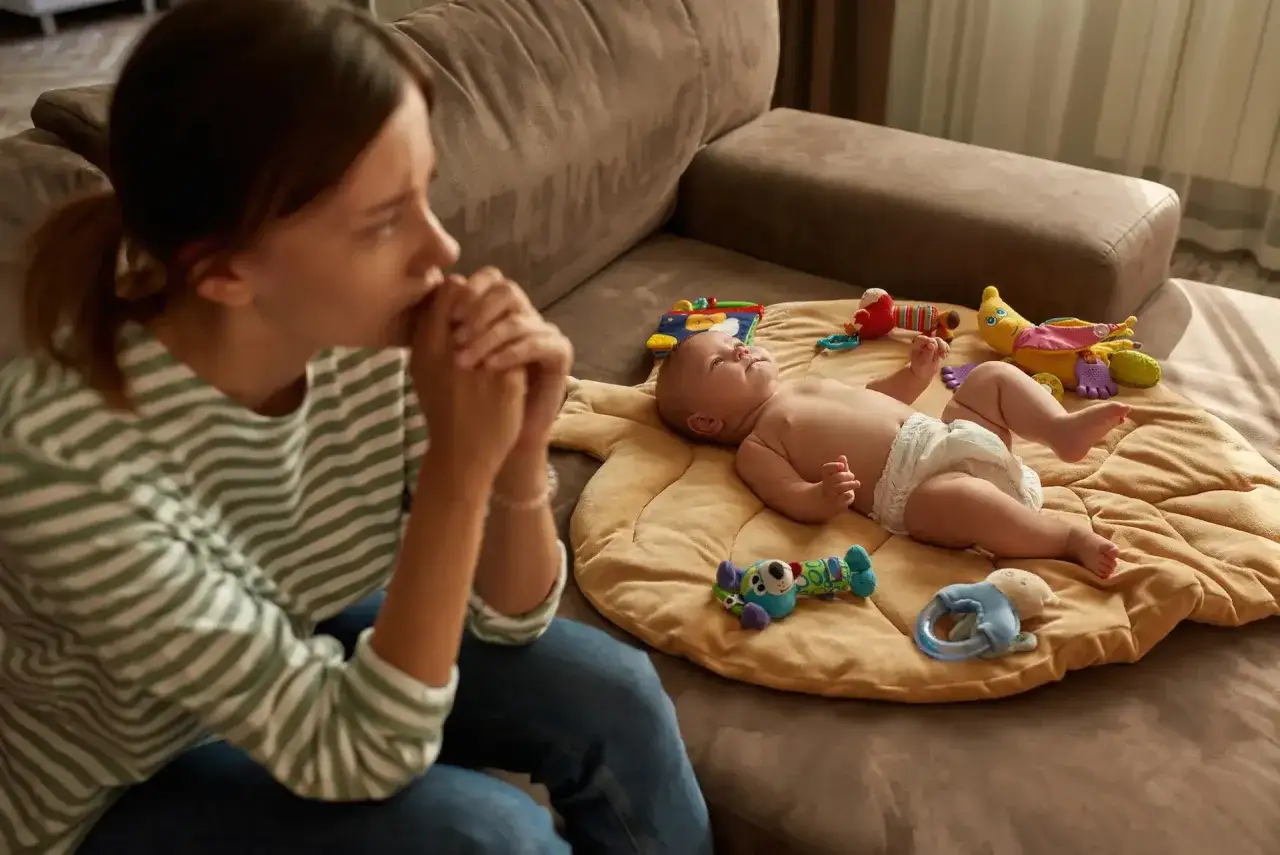 Giovane madre con le mani giunte, esausta, accanto al suo bambino che gioca. Il burnout genitoriale &egrave; palpabile.