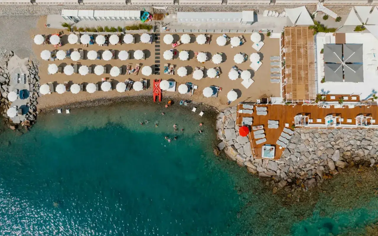 Vue a&eacute;rienne d'une **imperia plage** anim&eacute;e avec parasols blancs, baigneurs dans la mer turquoise et une terrasse en bois avec jacuzzi.