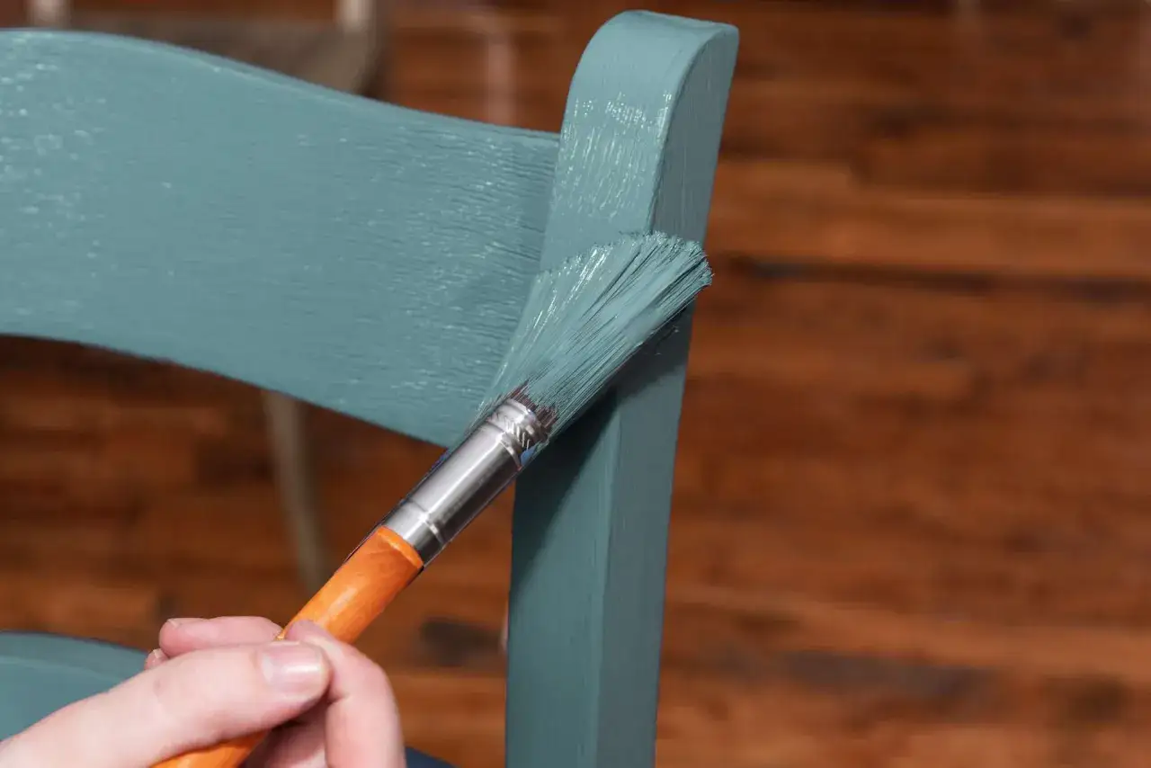 Mano pintando una silla de madera de azul verdoso con una brocha.