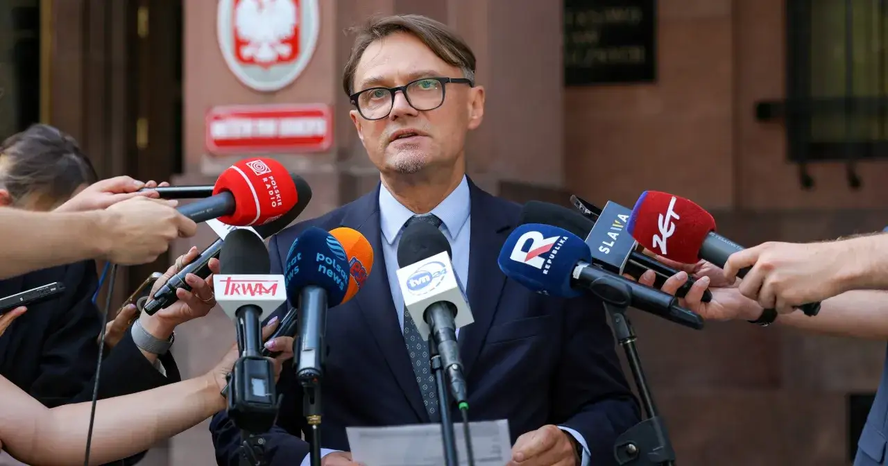 Mężczyzna w okularach, otoczony przez mikrofony różnych stacji, w tym TVN24, udziela wywiadu.