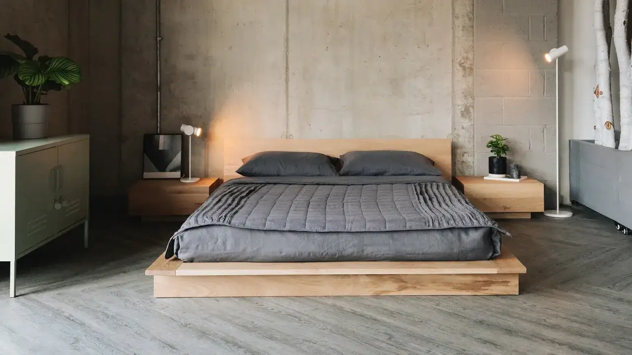 Ein Futonbett mit niedrigem Holzrahmen und grauer Bettw&auml;sche steht vor einer Betonwand. Minimalistische Nachttische und Lampen erg&auml;nzen den modernen Look.