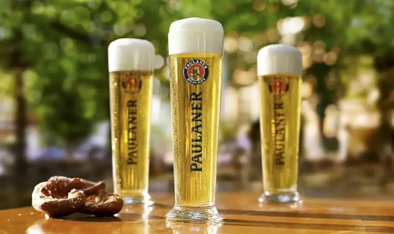 Tre bicchieri di birra hefe Paulaner con schiuma bianca e un pretzel salato su un tavolo di legno all'aperto.