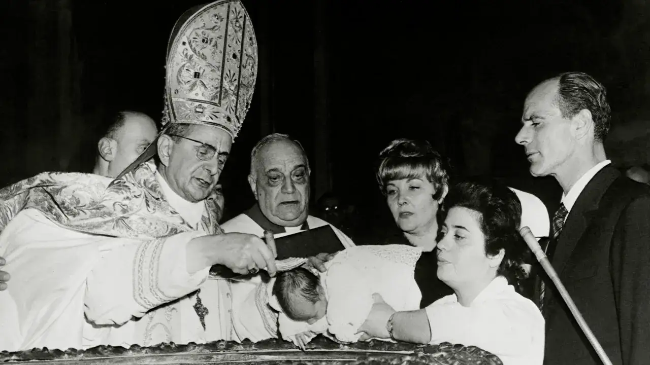 Papst Paul VI. tauft ein Baby. Eine Frau hält das Kind, während ein Mann daneben steht.