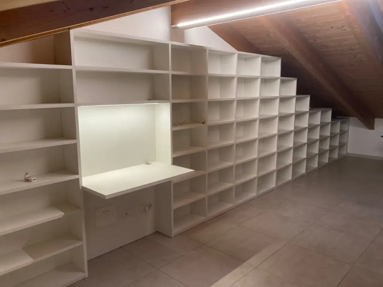 Ampia libreria mansarda bianca, con scrivania integrata e illuminazione a LED, perfetta per organizzare libri e oggetti in uno spazio sottotetto.
