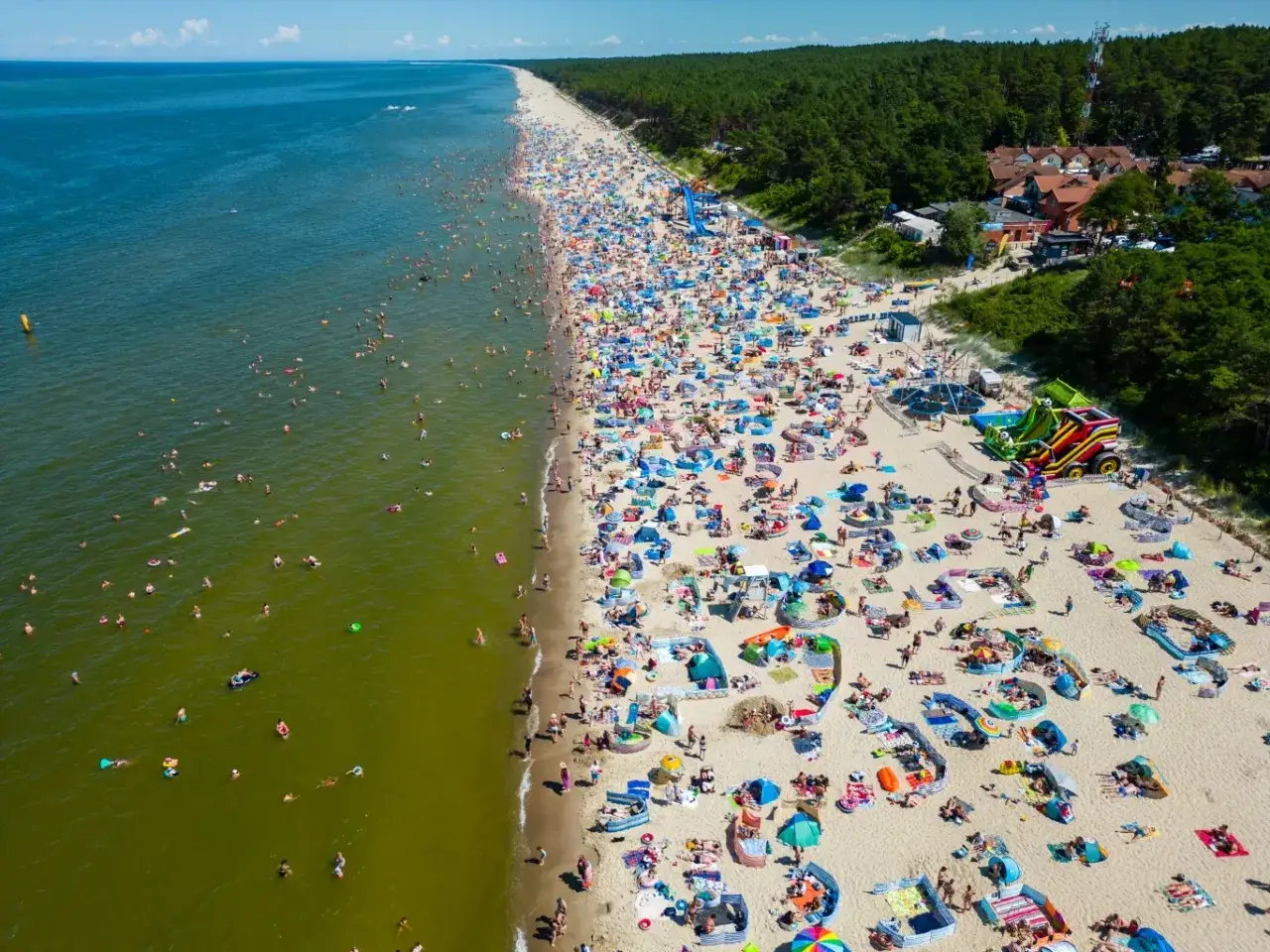 Tłumy ludzi na plaży, woda i las. Idealny **długi weekend sierpień** na wypoczynek nad morzem.