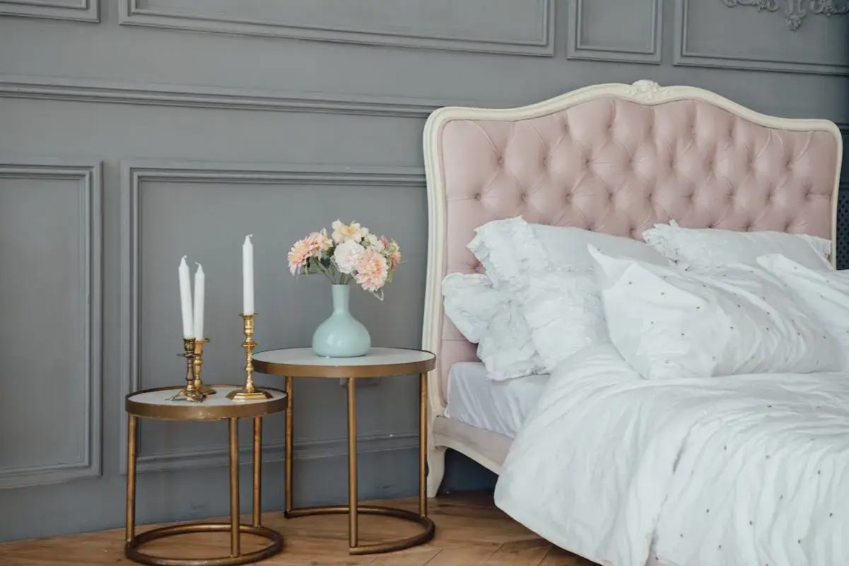 Sypialnia w stylu shabby chic z różowym, pikowanym zagłówkiem, białym pościelą i wazonem z kwiatami na stoliku.