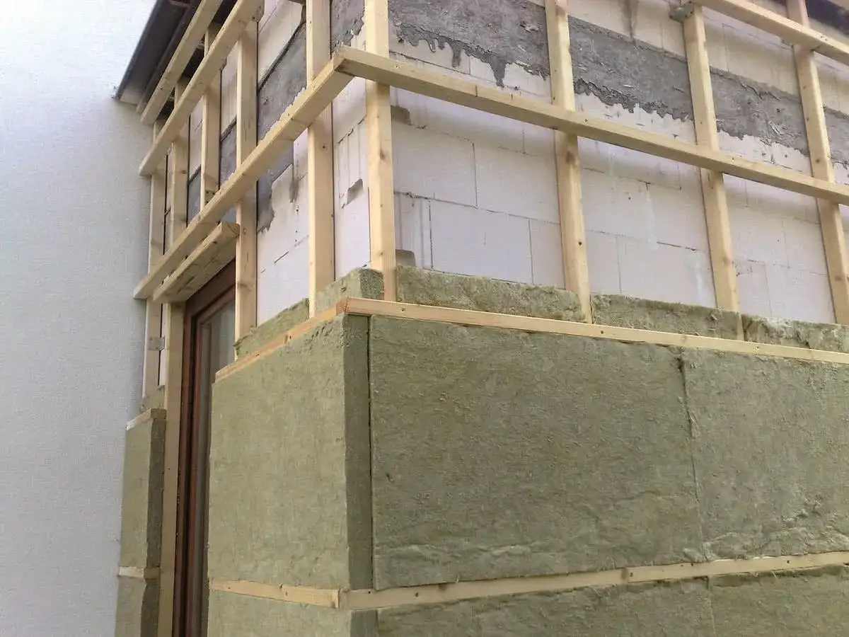 Montaż wełny mineralnej na ścianie zewnętrznej budynku. Grube warstwy izolacji są mocowane do drewnianej konstrukcji.