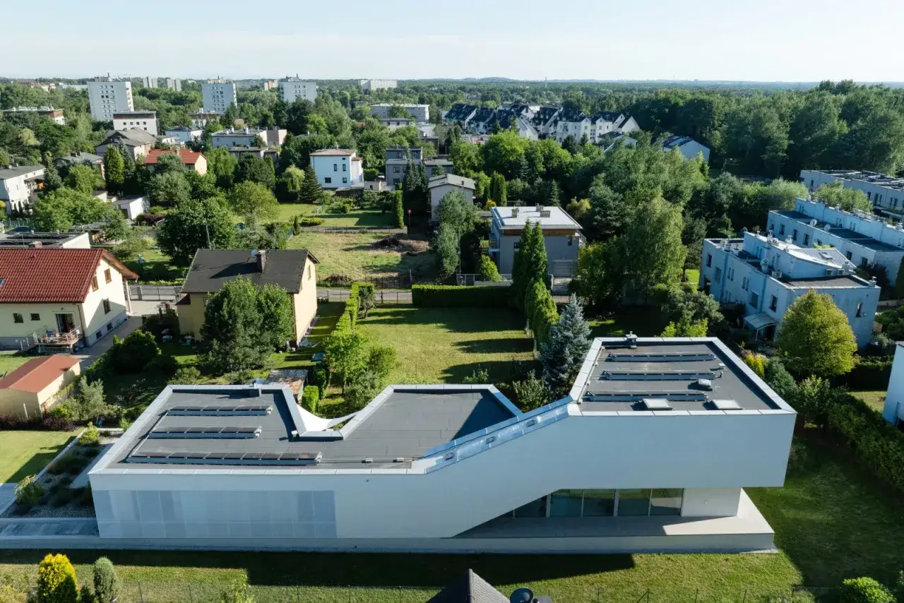 Nowoczesna bryła domu z płaskim dachem i panelami słonecznymi, przykład współczesnej architektury. W tle inne budynki i zieleń.