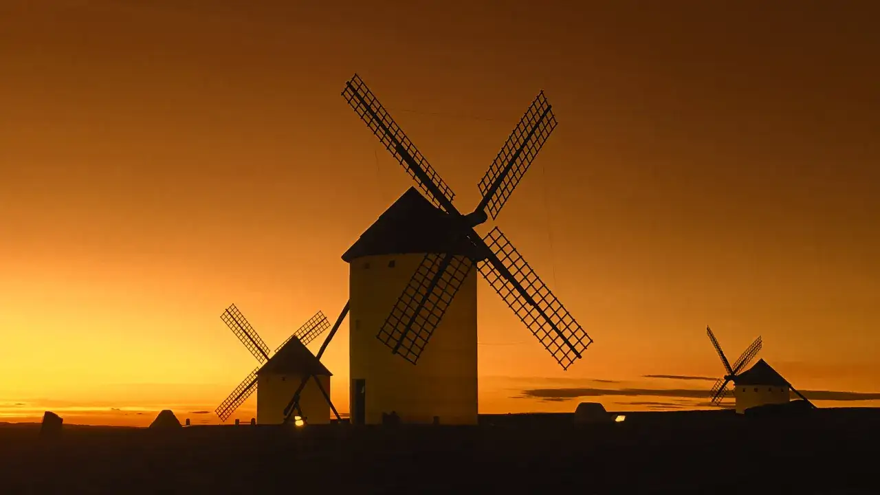 Molinos de viento silueteados contra un atardecer anaranjado, evocando la ruta del Quijote.