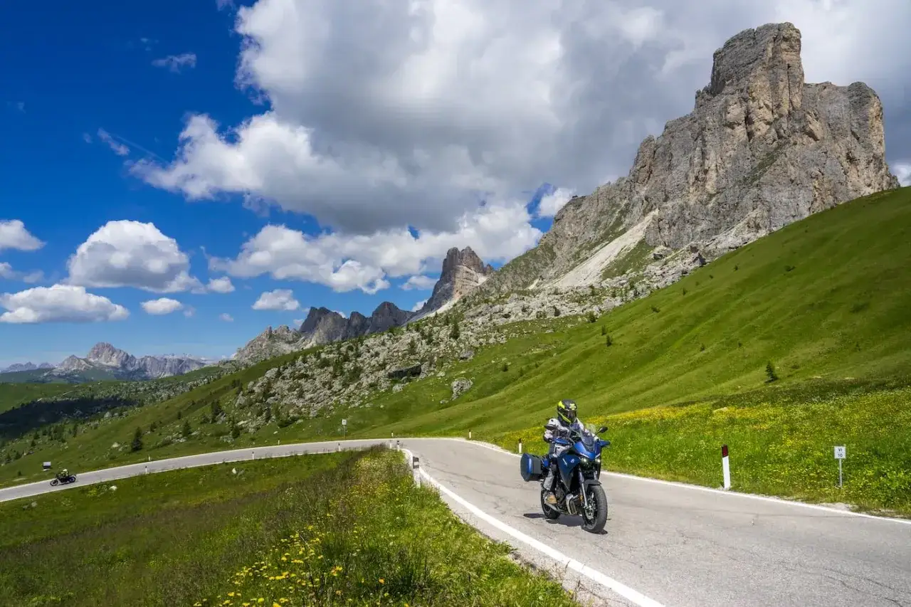 Motociclista su una strada panoramica in Veneto, tra verdi pendii e imponenti vette rocciose sotto un cielo azzurro con nuvole bianche.