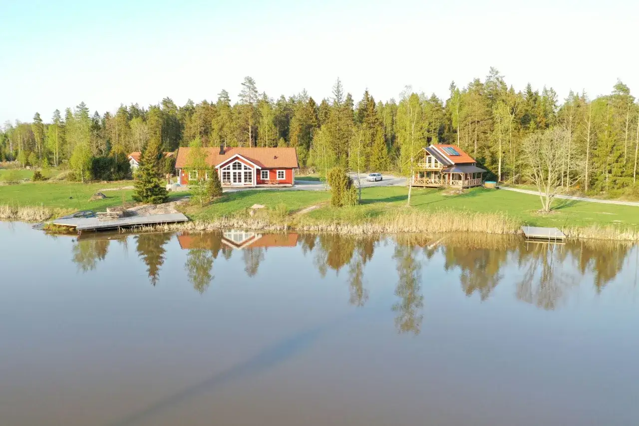 Två röda hus, en större med stora fönster och en mindre med veranda, ligger vid en sjö. En **gula stugan** syns inte, men det är en vacker vy.