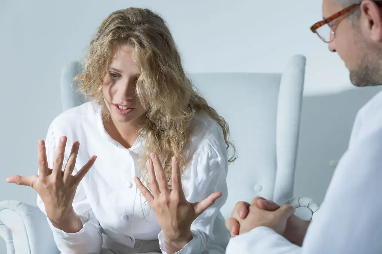 Kobieta z burzą blond włosów, w białej koszuli, gestykuluje podczas rozmowy z lekarzem. Może to być sesja terapeutyczna, podczas której omawiane jest leczenie, np. klozapina.